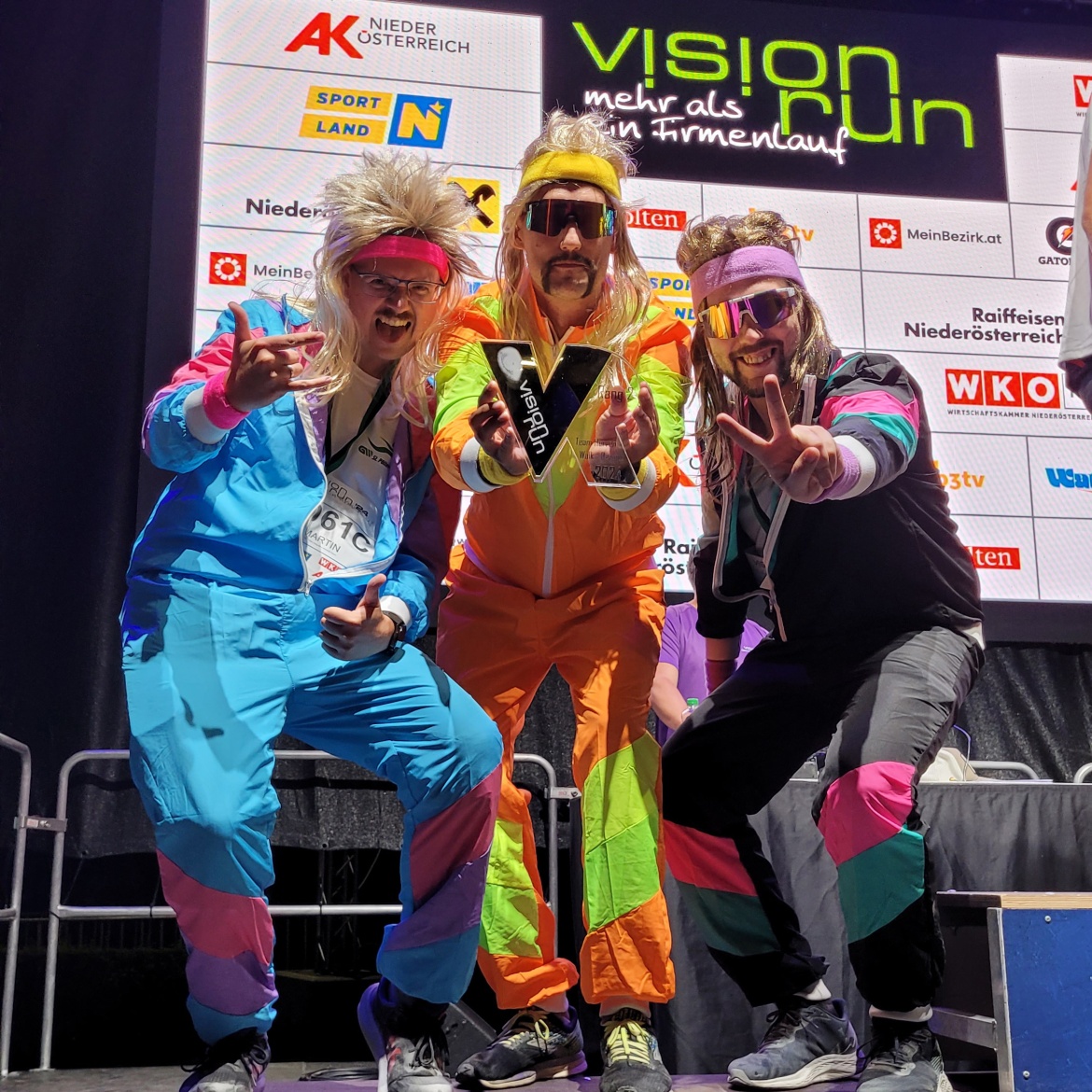 Gruppenfoto des 3er Männerteams, sie haben den zweiten Platz erlangt und halten ihren Pokal in den Händen. Alle drei sind haben lange Perücken und bunte Trainingsanzüge an.