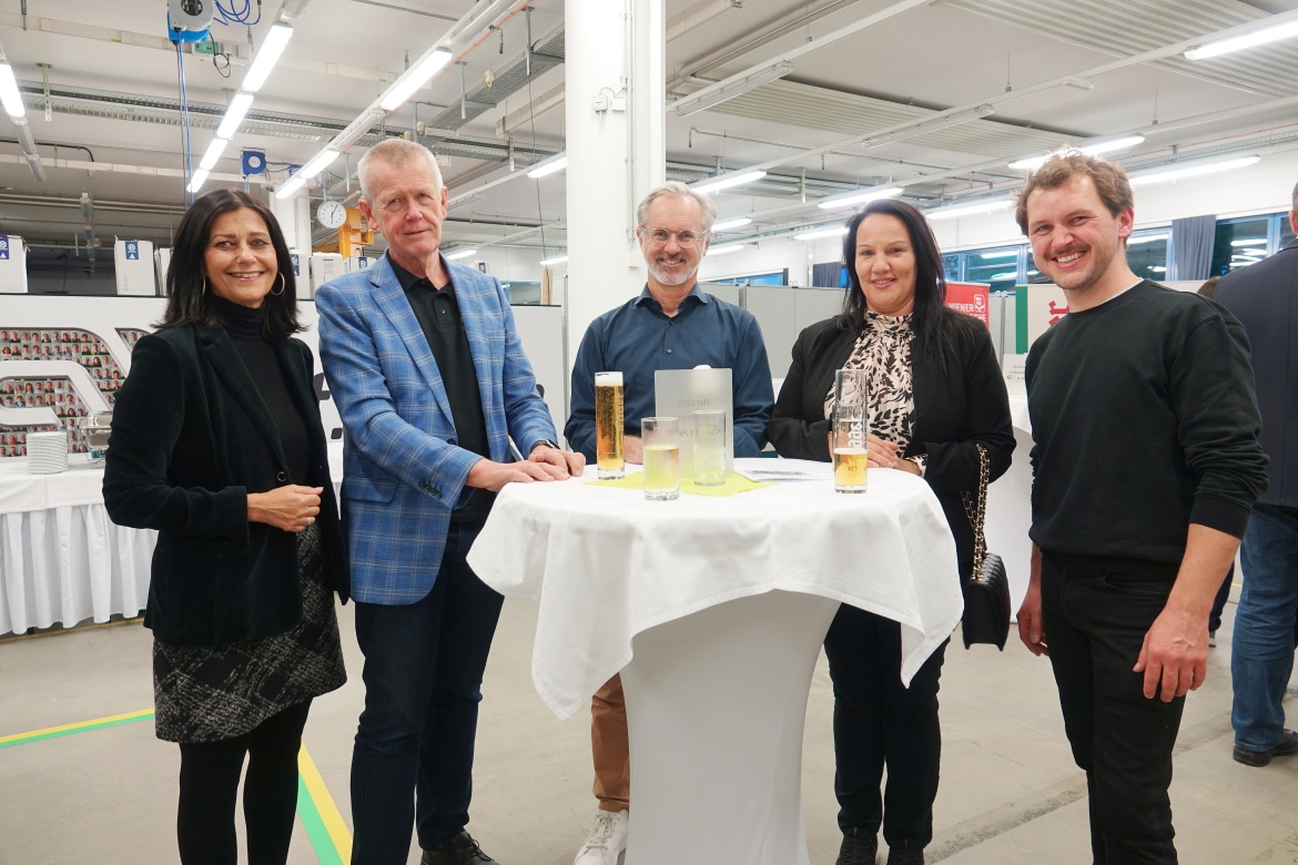 Eine Gruppe von Menschen steht um einen Stehtisch mit Getränken in der Hand